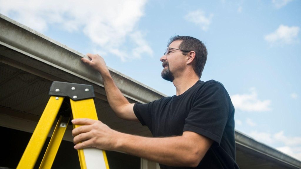 Man on a ladder inspecting a house gutter against a blue sky background.