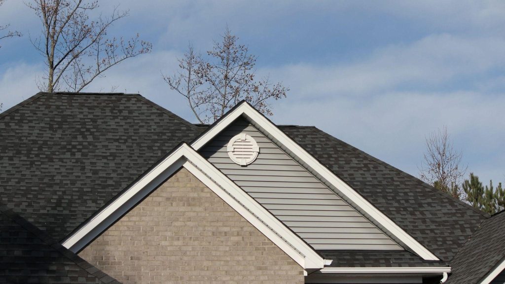 Close-up of a house roof with dark shingles, a gable vent, and a brick facade against a blue sky.