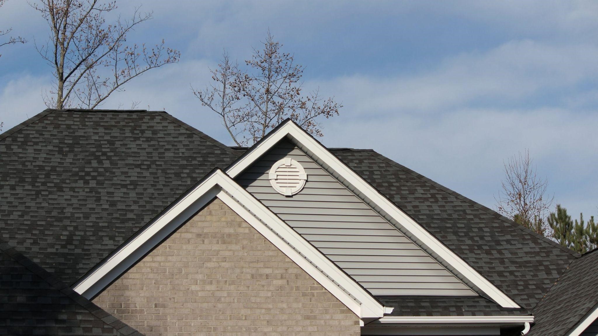 Close-up of a house roof with dark shingles, a gable vent, and a brick facade against a blue sky.