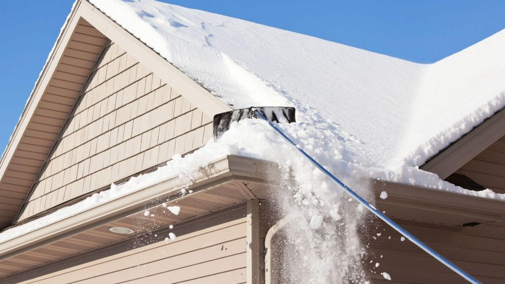 A roof rake removes snow from the edge of a house’s roof, with fresh snow falling over the gutter against a clear blue sky